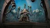 A detailed miniature figure stands prominently in the foreground, featuring a character with a complex helmet and multiple mechanical appendages extending from its back, each topped with small, glowing orbs. The figure is dressed in a dark, ornate robe, holding a device in one hand, while two other armored figures are partially visible in the background, suggesting a sci-fi setting. The scene is set against a moody, atmospheric backdrop that enhances the futuristic theme.