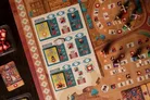A board game with a medieval village setting. Players place wooden devil and drummer miniatures on a grid. Cards show icons of devils, fireworks, and buildings with numbers and clock symbols. A player’s action board displays time slots, icons, and resource counters.