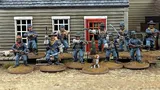 A group of ten detailed 28mm scale miniatures representing dismounted US Cavalry and Buffalo Soldiers stands in front of a wooden building with a red door. The miniatures are posed with various weapons, including rifles and a bugle, showcasing a mix of uniforms and accessories, such as scarves and hats. A small beagle and a cactus are also present among the figures, all mounted on 25mm round bases with a sandy terrain effect.