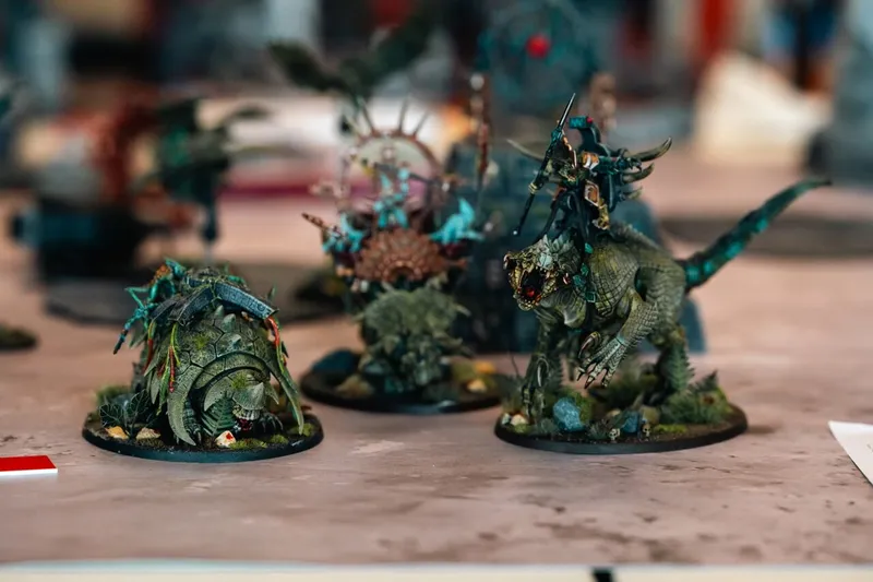 Three intricately painted tabletop miniatures are displayed on a flat surface. On the left, a large, armored creature resembling a tortoise is adorned with foliage and small details. In the center, a towering figure with a spiked shell and vibrant colors stands out, while on the right, a fierce, reptilian creature with a rider brandishing a weapon completes the trio. The background is slightly blurred, emphasizing the detailed craftsmanship of the miniatures.