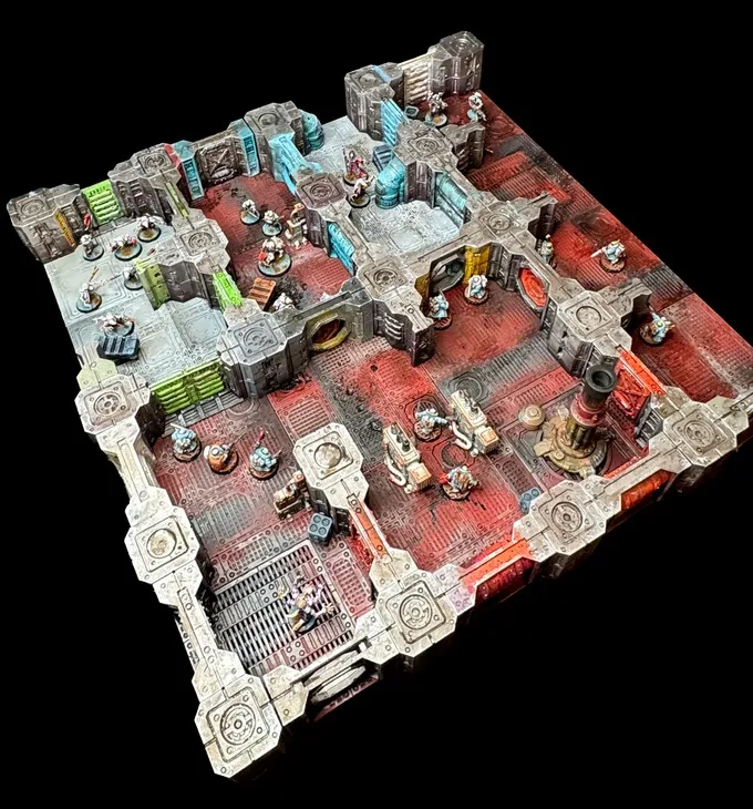 A detailed modular sci-fi terrain setup features a sprawling layout with various interconnected rooms and corridors, showcasing a mix of industrial and futuristic design elements. The terrain is painted in shades of gray, red, and green, with intricate textures and details, including pipes, machinery, and storage units. Miniature figures are positioned throughout the scene, suggesting a skirmish or exploration scenario within a derelict space environment.