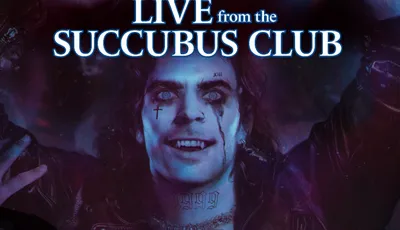 A vampire with pale skin and striking blue eyes is depicted in a dark, atmospheric setting, wearing a leather jacket and showcasing numerous tattoos across his chest and neck. His expression is exuberant, with a wide smile and streaks of dark makeup running down his face, suggesting a rock star persona. The background features a blend of purple and blue hues, enhancing the club-like ambiance, while the title "LIVE from the SUCCUBUS CLUB" is prominently displayed at the top.