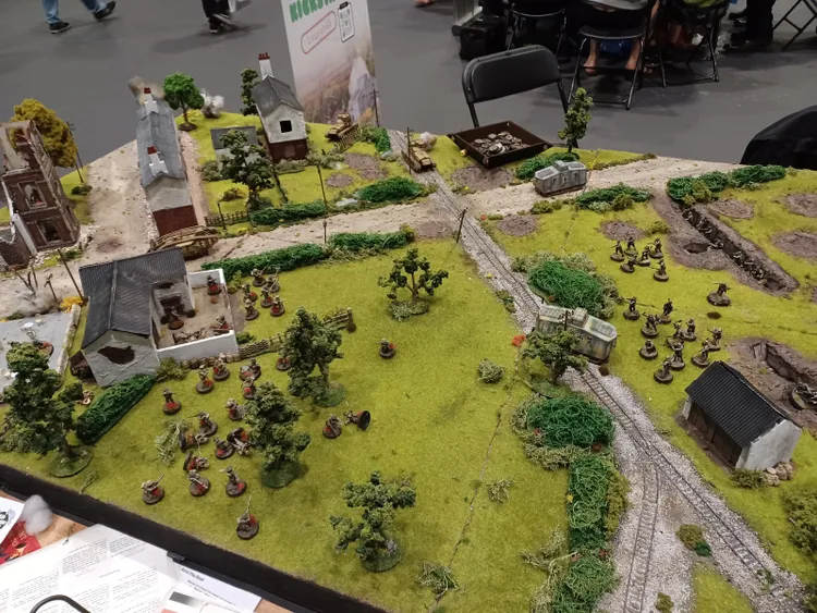 A detailed tabletop diorama depicting a World War I battlefield, featuring a lush green landscape with scattered trees, buildings, and a dirt road. Miniature soldiers from both the US Marine Corps and the German army are positioned throughout the scene, with some engaging in combat. The table also includes model vehicles and terrain features, such as a railway track and various structures, creating a dynamic and immersive setting for gameplay.