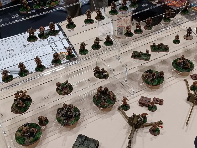A display of miniature soldiers arranged on clear acrylic stands, showcasing a variety of poses and equipment. The figures are painted in earthy tones, standing on circular bases with green grass tufts, suggesting a military theme. In the foreground, there are larger bases featuring groups of soldiers and weaponry, while smaller figures are positioned on the upper levels of the display.
