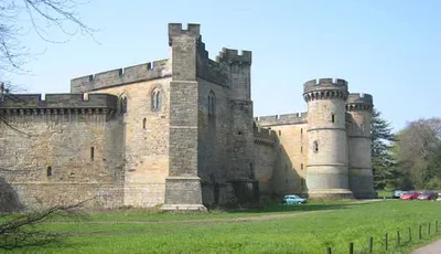 A large stone castle with multiple towers and thick walls, set against a clear blue sky. The castle features a mix of architectural styles, with rounded towers and crenellated parapets. In the foreground, a grassy area is bordered by a low fence, and a few cars are parked nearby.
