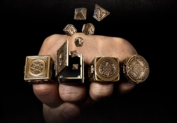 Four fantasy-themed rings are displayed on a hand, each featuring intricate designs and a hidden compartment for miniature RPG dice. The rings, crafted from brass with an aged finish, include motifs like a skull and various runes. Several polyhedral dice are shown emerging from one of the rings, highlighting their unique function as wearable dice vaults.