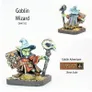 A detailed miniature of a Goblin Wizard stands prominently, showcasing vibrant colors and intricate features. The wizard wears a blue pointed hat and a flowing dark cloak, holding a staff topped with a skull in one hand and a spellbook in the other. The base is adorned with tufts of grass, emphasizing the 28mm scale of this character from the Goblin Adventurers range by Alternative Armies.