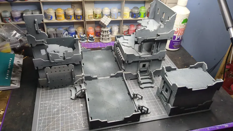 A partially assembled gray plastic model of a multi-level ruined building with detailed walls, staircases, and platforms, placed on a green cutting mat with grid lines. In the background, shelves hold numerous paint pots with labels such as "Citadel," "Vallejo," and "Army Painter," alongside small unpainted miniatures and a white spray bottle labeled "5 MIN SUPER FAST PV."