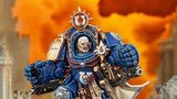 A detailed miniature of Marneus Calgar, a prominent character from the Warhammer 40,000 universe, is depicted in a striking pose. He wears ornate blue power armor adorned with gold embellishments, featuring a prominent Ultramarines symbol on his chest and a skull motif. The background is a dramatic orange, suggesting a fiery battlefield, enhancing the intensity of the scene.