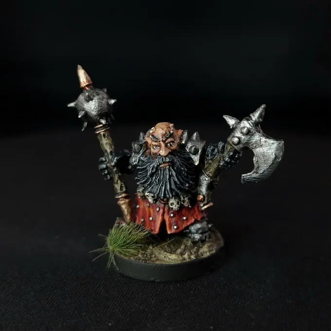 A painted miniature figure of a bearded dwarf-like warrior with dark skin, wearing a red and black outfit with metallic armor details. The figure holds a large double-headed axe with spiked ends in one hand and a spiked mace in the other. The base has small patches of green grass-like texture. The background is black.