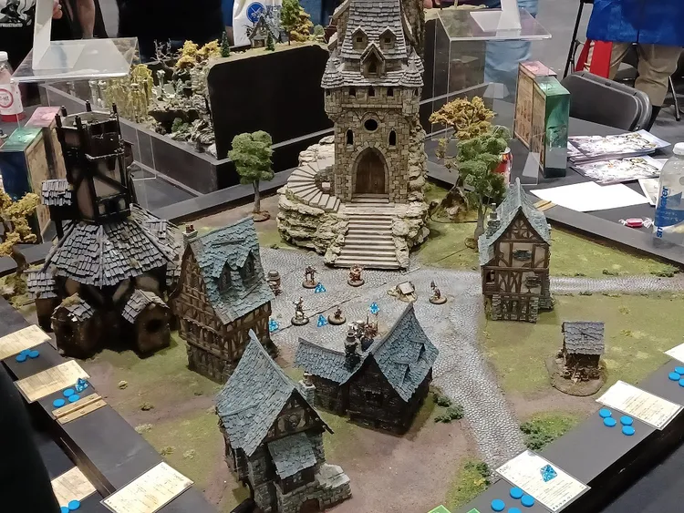 A detailed tabletop gaming scene features a variety of intricately designed buildings, including a tall stone tower with a steep roof and a winding staircase, surrounded by smaller houses with textured roofs and wooden facades. In the center, several miniatures are positioned on a cobblestone path, with blue tokens scattered around them, suggesting an active game in progress. The backdrop includes lush greenery and additional terrain elements, enhancing the immersive atmosphere of the display.