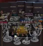 A detailed tabletop gaming scene showcases the "Elden Ring: The Board Game" campaign materials spread across a dark surface. Prominent in the background are three large game boxes labeled "Elden Ring: The Board Game," alongside various decks of cards and tokens. The foreground features numerous miniatures, including characters and creatures, arranged on a grid-based game board, highlighting the game's cooperative play and combat mechanics.