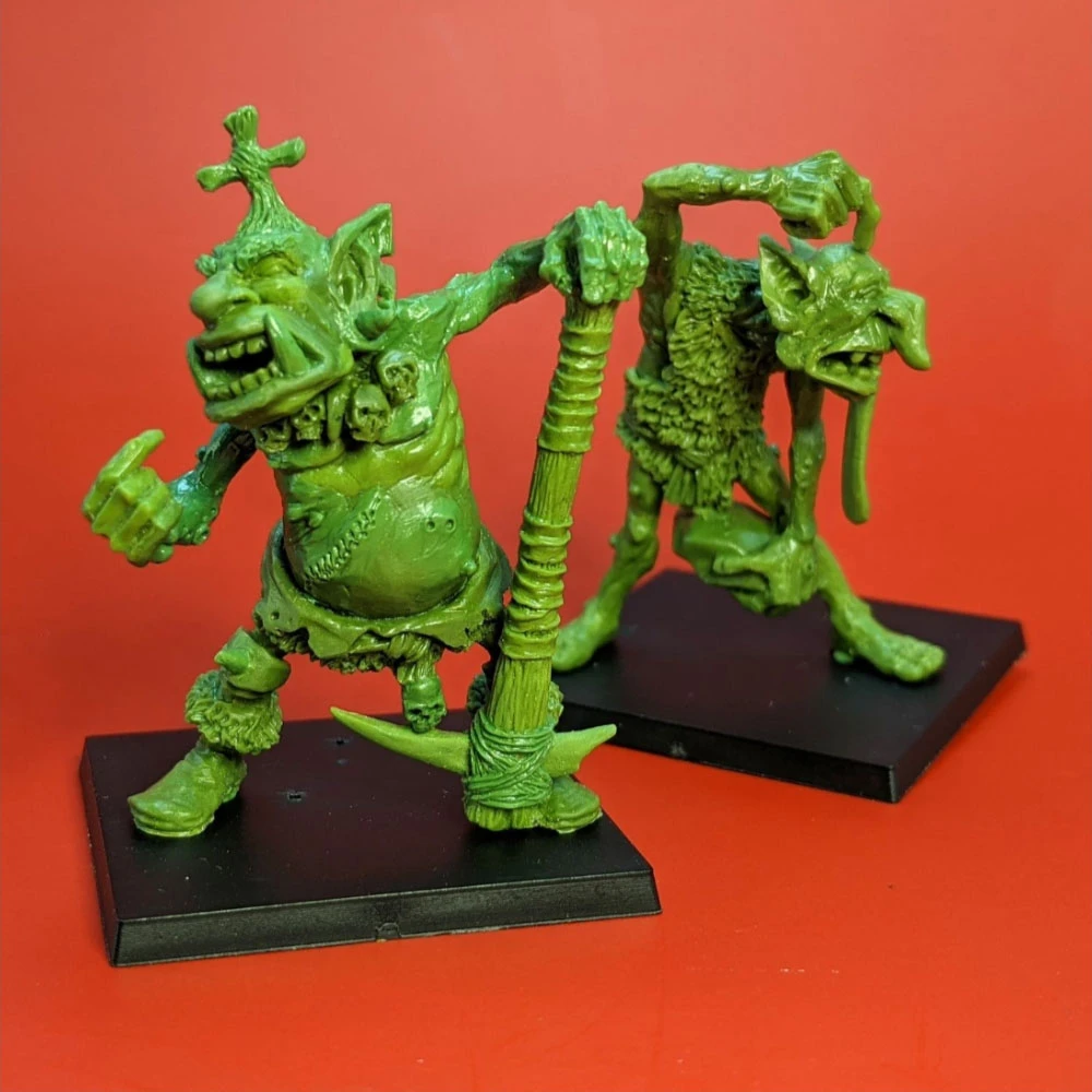 Two green hill troll miniatures stand on black bases against a bright red background. On the left, Thoughtful Grog is depicted with a broad grin, holding a large club, and adorned with various tribal elements. To the right, Grobna the Maw has a more sinister expression, featuring a hunched posture and a grotesque face, with a long, protruding tongue. Both figures showcase intricate details typical of fantasy miniatures.
