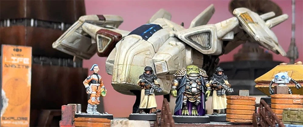 A detailed scene features a futuristic spaceship with a sleek design, prominently displayed in the background. In the foreground, several 28mm scale miniatures are arranged, including a female pilot in an orange jumpsuit, two armed security officers in dark uniforms, a large alien figure adorned in ornate armor, and a small robot. The setting includes wooden crates, enhancing the sci-fi atmosphere.