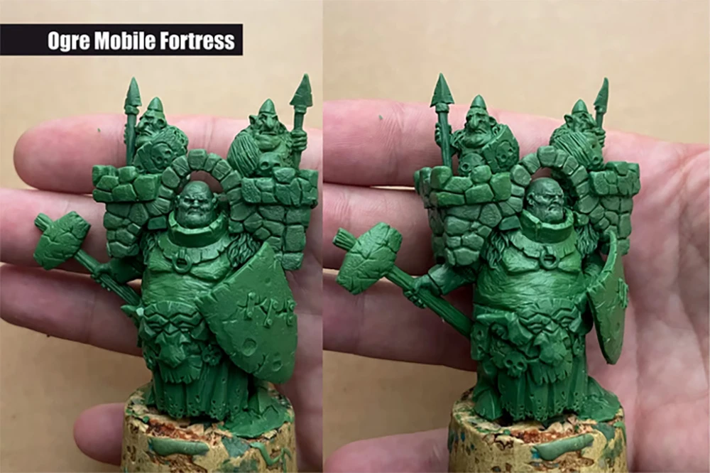 A detailed green sculpt of an "Ogre Mobile Fortress" is held in a person's hand, showcasing a large, rotund ogre character adorned with armor and a shield. The ogre is flanked by two smaller figures, likely goblins, peeking over a stone wall structure on the ogre's back. The sculpt features intricate details, including facial expressions and textures, emphasizing the whimsical and mischievous nature of the characters.