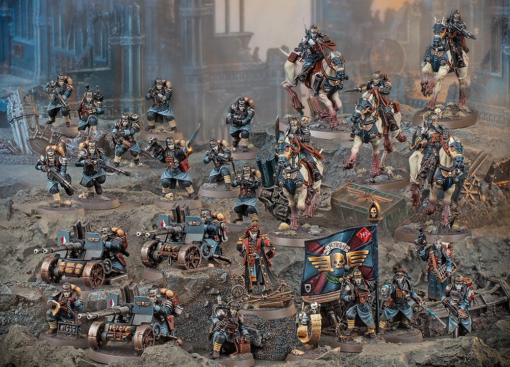 A detailed display of the Death Korps of Krieg models features a variety of miniatures, including ten troopers, a Command Squad, a Heavy Weapons Squad, and five cavalry riders on horses. The models are painted in muted tones of blue and gray, with some adorned in red accents, set against a rocky terrain backdrop. A flag bearing the Death Korps insignia and a few heavy weapon emplacements are also visible, emphasizing their military theme.