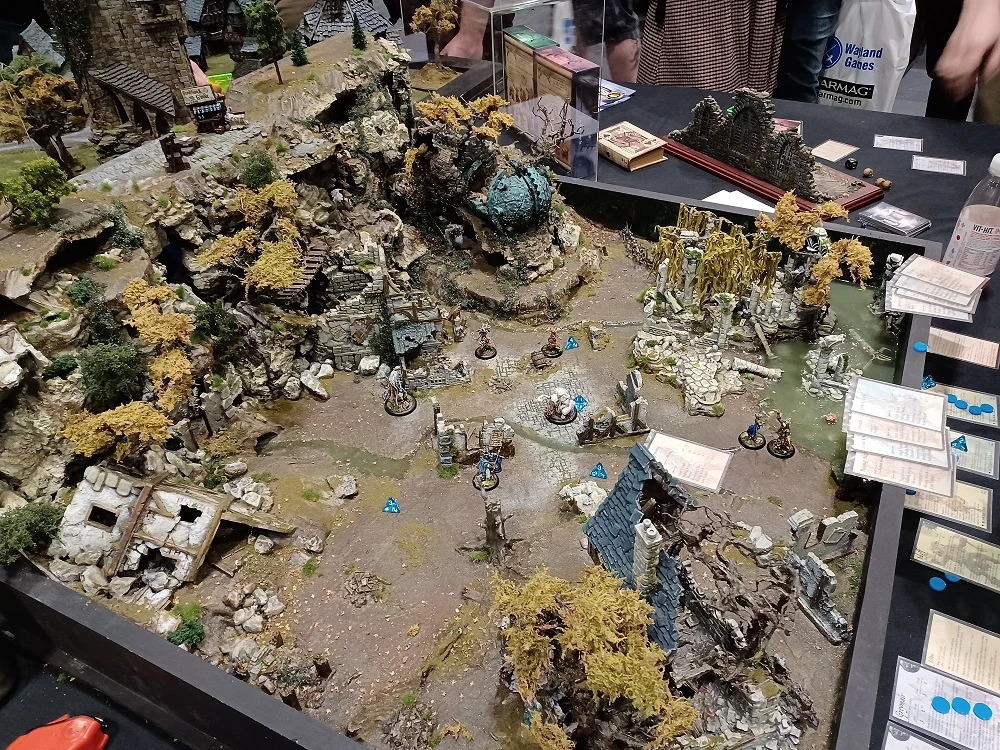 A detailed tabletop gaming scene showcases a rugged terrain with rocky outcrops, scattered ruins, and vibrant foliage in autumnal colors. Miniatures are positioned throughout the landscape, engaging in a skirmish, with some figures standing on stone pathways and others near dilapidated structures. The backdrop features a mix of natural elements and remnants of a once-thriving settlement, creating an immersive environment for gameplay.