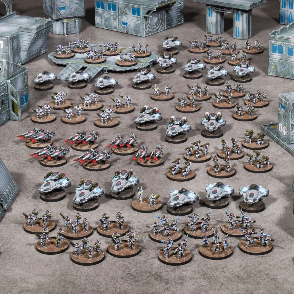 A large array of 10mm-scale miniatures is displayed on a tabletop, showcasing a sci-fi wargaming scene. The scene features various types of vehicles and infantry, including sleek hovercraft and armored soldiers, all set against a backdrop of modular, futuristic buildings. The miniatures are arranged in organized formations, highlighting the two distinct armies, with a mix of colors primarily in shades of gray and metallic tones.