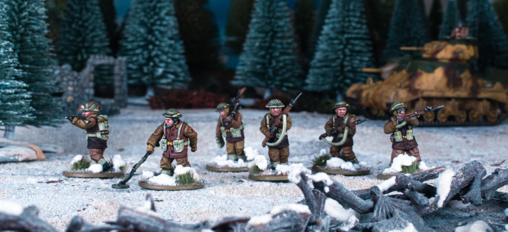 A group of six multi-part British and Canadian winter infantry figures stands on a snowy terrain, surrounded by evergreen trees and a tank in the background. Each figure is dressed in warm winter gear, showcasing a variety of poses and equipped with different weapons, including rifles and sidearms. The scene captures the essence of a winter battlefield, with patches of snow and scattered debris enhancing the atmosphere.