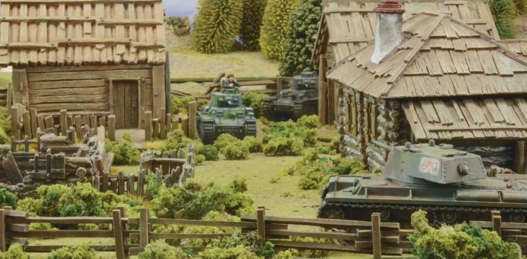 A detailed tabletop scene features several miniature tanks amidst a rural setting, including a wooden cabin and lush greenery. In the foreground, a tank with the number "600" is positioned near a wooden fence, while another tank is seen further back with a crew member visible. The background includes trees and additional structures, creating a vibrant battlefield atmosphere.