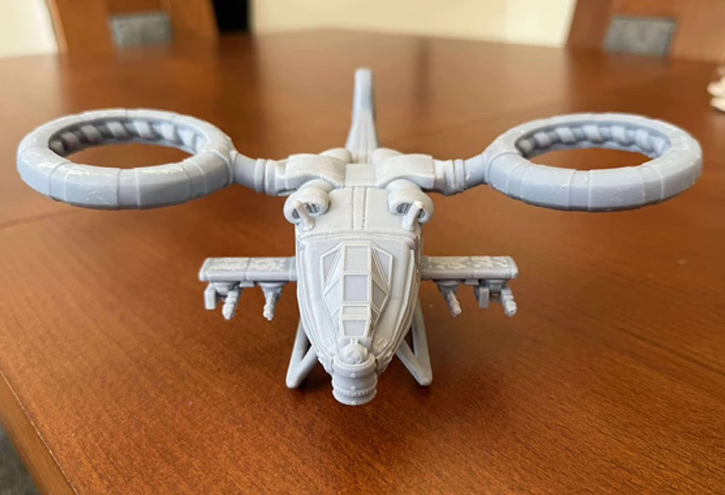 A detailed model of the LARC 2, a sci-fi utility VTOL, is displayed on a wooden table. The model features a streamlined design with a pointed nose, twin anti-grav rotors, and various weapon hardpoints on the sides. The overall color is a light gray, showcasing intricate details such as cockpit windows and structural elements.