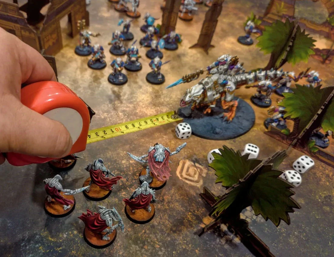 A tabletop gaming scene features a vibrant battlefield with various miniatures positioned across a textured gaming mat. In the foreground, a player is using a measuring tape to gauge distance between a group of robotic figures in red capes and a large, dinosaur-like creature with a rider, which stands on a circular base. Surrounding them are additional miniatures, terrain pieces, and several white dice scattered on the ground, creating an immersive gaming environment.