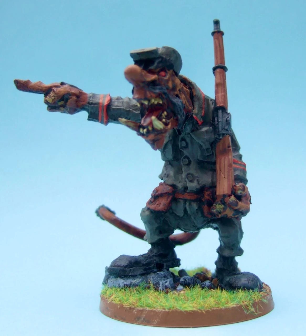 A detailed miniature of a humanoid figure, resembling a soldier, stands on a grassy base. The figure has a grotesque, exaggerated face with sharp teeth and a fierce expression, wearing a military uniform that appears tattered and worn. It is pointing forward with one hand while holding a rifle in the other, showcasing a dynamic pose that conveys aggression and readiness for battle. The background is a soft blue, emphasizing the figure's vibrant colors and intricate details.