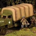 A military scene features a green WWII truck with a large, canvas-covered cargo area, displaying a white star insignia on its side. Surrounding the truck are several soldiers in period-appropriate uniforms, some aiming their weapons and others in various poses, set against a grassy terrain with trees in the background. The overall atmosphere conveys a sense of readiness and action typical of wartime scenarios.