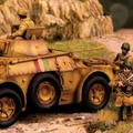 An Italian Autoblinda AB41/43 Armoured Car is prominently featured in the foreground, showcasing a weathered yellow and brown camouflage pattern. The vehicle has a turret with a soldier positioned on top, while several infantry soldiers in tan uniforms are scattered around it, engaged in various poses. The background includes rocky terrain and greenery, enhancing the military setting.
