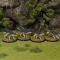 Four fantasy wolf miniatures are displayed on a grassy terrain, each mounted on a circular base. The wolves have detailed fur textures, featuring dark gray and brown coloring, with some showing dynamic poses as if in motion. In the background, there are small trees and rocky formations, enhancing the natural setting.