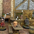 A detailed scene inside a fantasy tavern features several painted miniatures. In the foreground, a woman in a red skirt brandishes a sword, while a man in a blue tunic stands nearby. Behind the bar, a female bartender interacts with a cloaked figure, and various bottles and mugs are visible on the shelves. The tavern's rustic wooden furniture and stone walls create an immersive atmosphere.