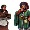 A miniature figure of a woman in a brown dress and white apron holds a tray filled with loaves of bread, smiling as she stands on the left. Beside her, a man in a colorful green cloak and red hat raises a cup to his lips, wearing a sly expression. Both figures are intricately detailed, showcasing the vibrant colors and textures typical of fantasy tavern patrons.