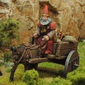 A miniature figure of a gnome with a long beard and a red pointed hat sits atop a wooden cart pulled by a dark brown donkey. The cart is laden with various items, including sacks and a small barrel, and is set against a backdrop of rocky terrain and green foliage. The scene captures a whimsical, fantasy-inspired atmosphere, perfect for a tabletop RPG setting.
