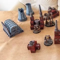 A collection of Epic scale terrain pieces suitable for games like Adeptus Titanicus and Battletech is displayed on a tan surface. The terrain includes various structures, such as a large, blue-gray building with pipes, several red industrial buildings, and tall smokestacks. Among the terrain, two detailed miniatures resembling mechs are positioned, adding to the scene's dynamic and futuristic atmosphere.