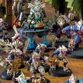 A dynamic scene featuring a variety of intricately designed miniatures from the Saurian Starhost army, showcasing colorful lizardmen in futuristic armor, armed with weapons. In the center, a larger figure sits on a throne-like structure, adorned with ornate details, while smaller lizardmen flank it, some wielding guns and others displaying fierce expressions. The background includes hints of lush foliage and a textured ground, enhancing the sci-fi atmosphere of the setting.