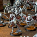 A detailed scene showcases a variety of intricately designed robot miniatures, each with a metallic silver and blue color scheme, set against a textured brown base. The figures include humanoid robots with flowing capes, armed with futuristic weapons, and larger mechanical constructs in the background. The composition highlights the diversity of the robot legions, emphasizing their advanced technology and battle-ready stance.