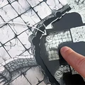 A close-up view of a modular mapping system for tabletop gaming, featuring a detailed, grimdark-style map with various sections labeled as Q2, R2, R4, R5, and R6. Two fingers are positioned on the map, indicating specific areas, while an arrow points from Q2 to R2, suggesting a connection or pathway. The textured surface of the map includes intricate stone patterns, enhancing the fantasy theme.