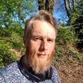 A man with a light beard and short hair stands outdoors, surrounded by lush green trees and foliage. He wears a patterned shirt featuring floral designs and has headphones around his neck. The sunlight filters through the leaves, creating a bright and inviting atmosphere.