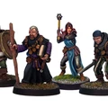 A group of four detailed miniatures representing classic adventurer character classes. On the left, a knight in armor holds a shield, followed by a robed figure with a book and staff. The third figure is a female illusionist in a blue dress with a staff, and on the right, a rugged fighter brandishes a sword. Each miniature is mounted on a round base with grass detailing.