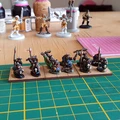 A collection of painted 10mm Orc miniatures is displayed on a green cutting mat, showcasing various poses and weapons. The Orcs are arranged on a wooden base, with some holding spears and shields, while others wield axes and a banner. In the background, partially visible models and tools suggest an active painting workspace, with bottles and a pink cutting mat adding to the scene's hobbyist atmosphere.