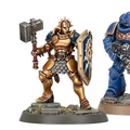 A detailed miniature of a Stormcast Eternals Liberator stands on the left, wielding a large hammer and a shield adorned with a lion emblem, showcasing a gold and blue color scheme. To the right, an Infernus Space Marine is depicted in blue armor, holding a weapon and featuring intricate detailing on its armor. Both miniatures are positioned on textured bases with grass and stone elements.