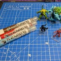 Three Playmarkers from AK are displayed on a blue cutting mat, featuring a grid pattern. The markers are in red, blue, and white, each labeled "PLAYMARKERS FOR TABLETOP GAMERS." In the foreground, two small miniatures are positioned on clear bases: a black creature and a red creature, while a larger green creature and a blue figure are visible in the background.