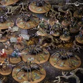 A detailed tabletop scene features various miniatures from the Middle-earth Strategy Battle Game, depicting a tense moment in Mirkwood. Prominent are several large spiders on circular bases, surrounded by a mix of characters including Bilbo, Thorin, and other dwarves, all positioned on a textured terrain with rocks and twisted roots. The dark, earthy colors of the setting enhance the atmosphere of the iconic "Flies and Spiders" chapter from The Hobbit.