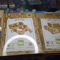 Two packages of TT Combat tabletop scenery are displayed on a work surface. The left package is labeled "Iron Labyrinth: Death Quadrant Complex," featuring a design of interconnected structures. The right package is labeled "Iron Labyrinth: Floors," showcasing a stack of storage tanks. In the background, various paint pots and additional terrain pieces are visible, indicating an active hobby workspace.