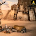 A detailed terrain setup features a sandy landscape with a large, weathered structure in the background, showcasing various mechanical elements and textures. In the foreground, a small, futuristic outpost with a rounded top is positioned next to a covered crate, while a small creature with blue wings and a scaly body rests nearby. The overall scene evokes a desolate yet intriguing atmosphere, suitable for tabletop gaming.