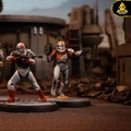 A trio of detailed miniature figures from the Delta Squad, featuring Clone Commandos Boss, Fixer, and Scorch, stands on circular bases. Each figure is uniquely painted, showcasing their distinct armor designs: Boss in gray with red accents, Fixer in gray with blue details, and Scorch in gray with orange highlights. The background depicts a desolate, industrial landscape, enhancing the battle-ready theme of the scene.
