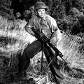 A black-and-white image features a soldier in a military uniform, wearing a helmet and a sergeant's insignia, crouched in a grassy field. He holds a machine gun with both hands, poised as if ready for action. The background consists of blurred greenery, suggesting a natural setting.