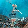 A dynamic scene features an Idoneth Deepkin warrior, the Ikon of the Sea, poised on a wave of water, wielding a curved sword and adorned with intricate armor and a decorative headdress. In the background, remnants of a shipwreck are visible, with splashes of water and a menacing sea creature emerging from the waves. The overall color palette includes vibrant blues and earthy tones, enhancing the aquatic theme of the model.
