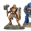 A golden-armored Stormcast Eternals Liberator stands on a rocky base, wielding a large hammer in one hand and a shield emblazoned with a sun motif in the other. Next to it, a blue-armored Infernus Space Marine holds a firearm, showcasing intricate details on both miniatures. The base features tufts of grass and rocky terrain, enhancing the overall display of these Warhammer figures.