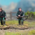 Two detailed miniatures of soldiers from the Sharpe’s Rifles series are displayed on textured bases, set against a blurred natural background. Both figures wear green uniforms with black crossbelts; one holds a rifle and has a serious expression, while the other appears to be in a more contemplative pose. The bases feature tufts of grass, enhancing the realistic terrain effect.